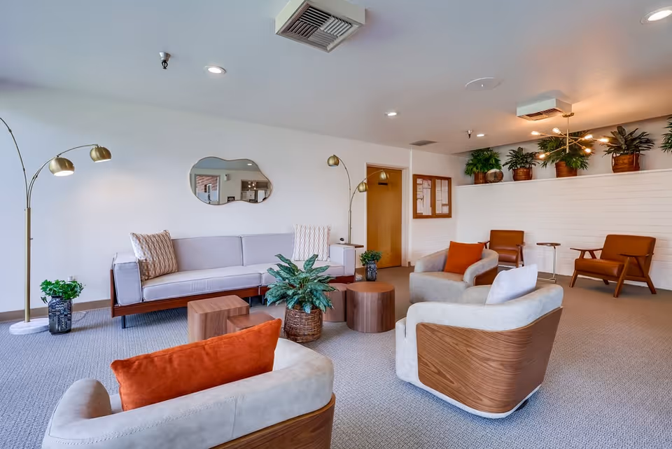 A well-lit nursing home common area with modern furniture including a light gray sofa, two beige armchairs with wooden accents, two brown armchairs, and several small wooden tables. The room features potted plants, a decorative mirror on the wall, and two gold floor lamps. The walls are white, and the carpet is light gray.