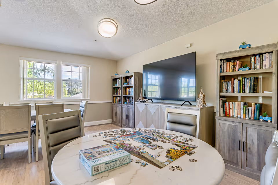 A bright room with a round table in the foreground featuring a partially completed jigsaw puzzle and a puzzle box. The room has light-colored walls and wood flooring. There are two wooden bookshelves filled with books and decorative items, a large flat-screen TV mounted on a cabinet, and a window letting in natural light. Several chairs are arranged around the tables.