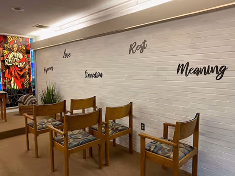 Interior room with wooden chairs arranged in rows facing a white brick wall. On the wall are words in black cursive font: Purpose, Love, Connection, Rest, and Meaning. A colorful stained glass window is visible on the left side of the room.