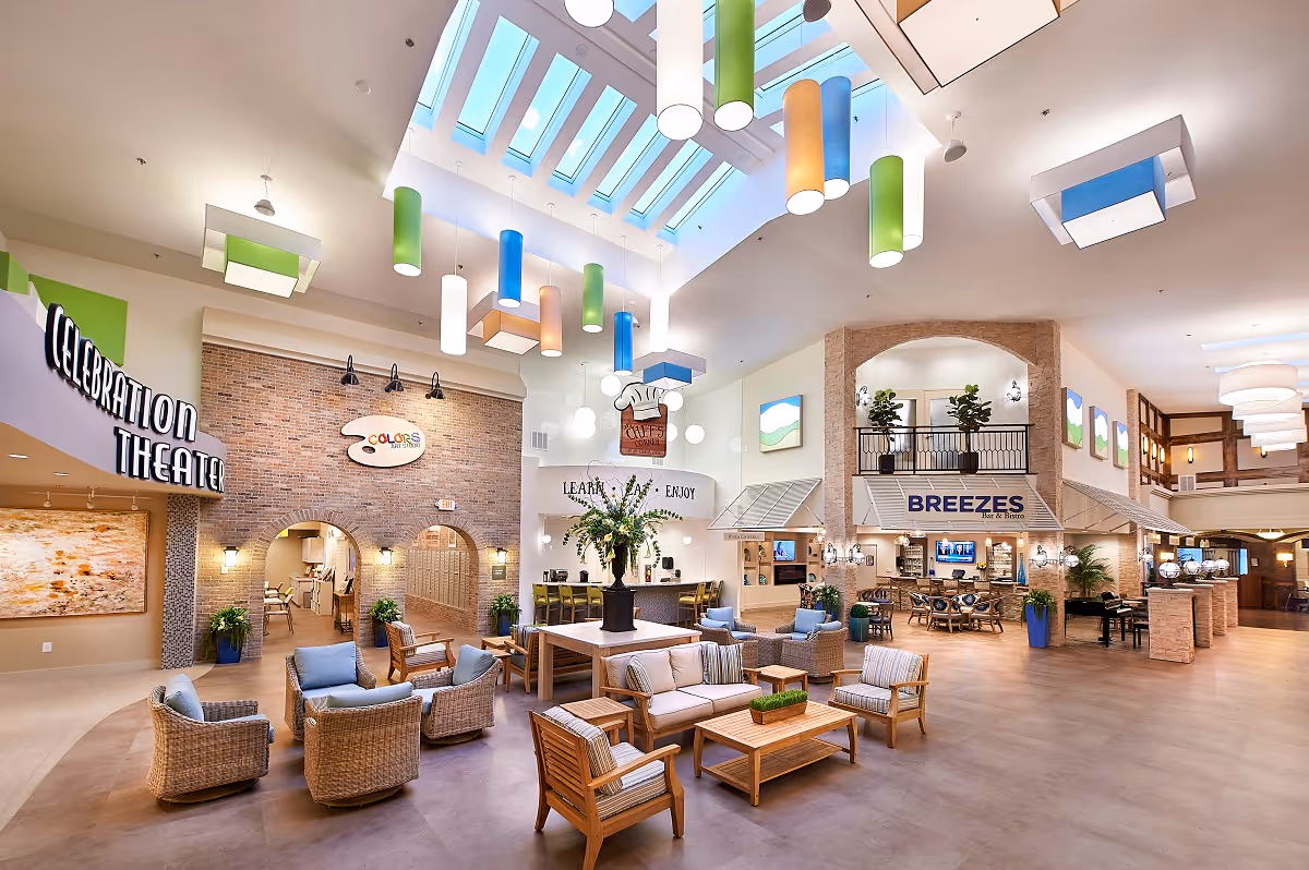 Spacious and brightly lit common area in a senior living facility featuring comfortable seating with wicker and wooden chairs and sofas arranged around coffee tables. The ceiling has large skylights and colorful hanging cylindrical lights. Visible signs include 'Celebration Theater', 'Colors Art Studio', and 'Breezes Bar & Bistro'. The area has a welcoming and modern design with plants and artwork on the walls.