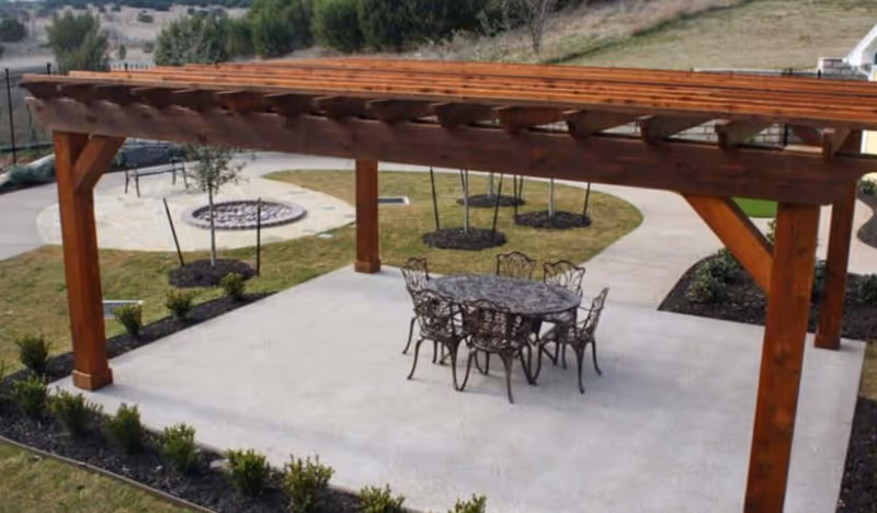 Wooden pergola over a concrete patio with a metal table and chairs and landscaped yard including a circular fire pit.