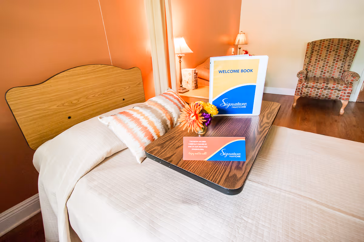 A neatly made bed with a wooden headboard in a room with peach-colored walls. On the bed is a wooden tray holding a welcome book labeled 'Signature HealthCARE' and a small card. There is a colorful flower arrangement on the tray. In the background, there is a patterned armchair and a side table with a lamp.