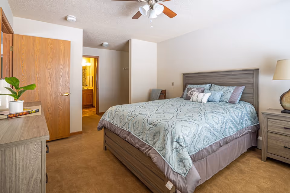 Furnished bedroom with a queen bed featuring a patterned comforter, wooden dresser and nightstand, and an open door to an adjacent bathroom.