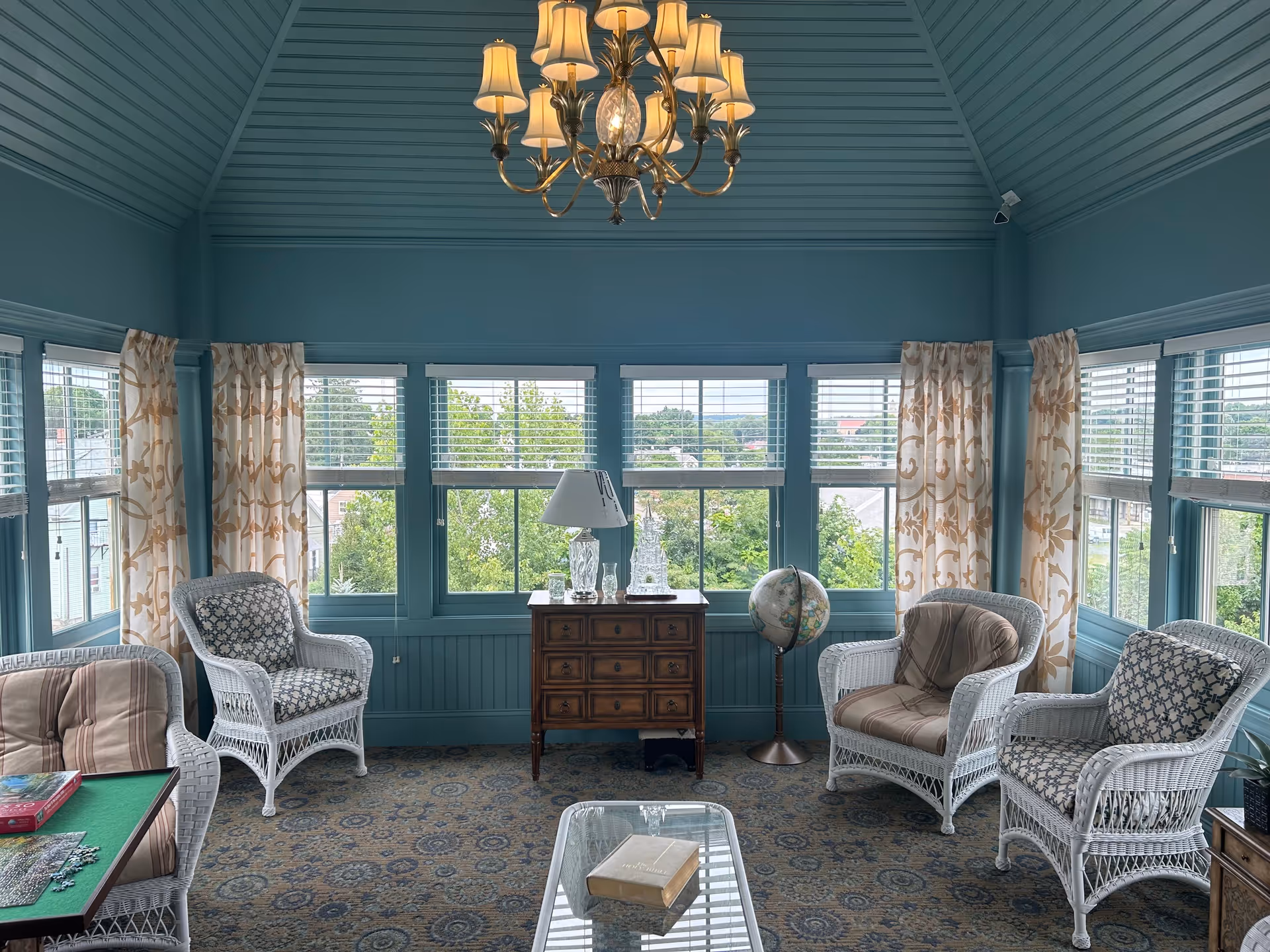 A cozy sitting room with teal walls and ceiling, featuring large windows with white blinds and patterned curtains. The room contains white wicker chairs with cushions, a wooden chest of drawers with a lamp and glass decor on top, a globe on a stand, and a glass coffee table with a book. A chandelier with multiple lampshades hangs from the ceiling.