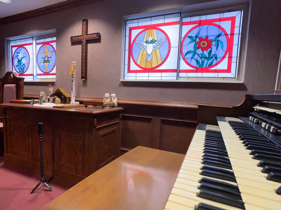 Interior of a chapel or prayer room with stained glass windows depicting religious symbols, a wooden cross on the wall, a wooden altar with religious figurines, and a keyboard organ in the foreground.