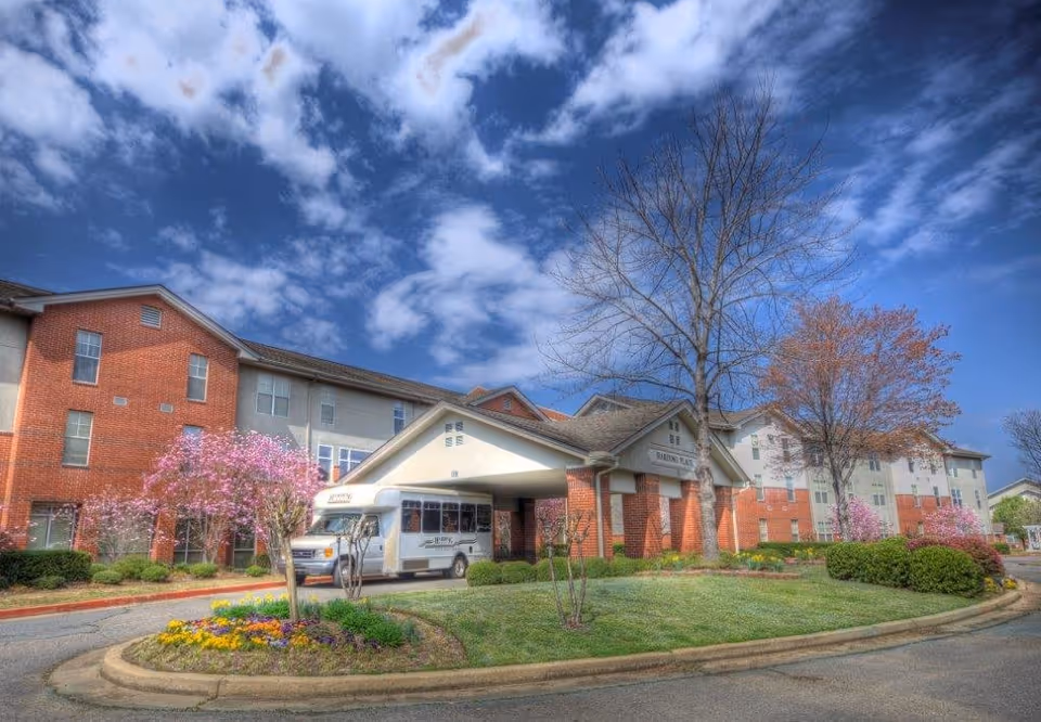 Exterior view of Harding Place Retirement facility showing a large multi-story building with red brick and beige siding. There is a covered entrance with a white shuttle bus parked underneath. The foreground features a landscaped roundabout with colorful flowers and small trees, some with pink blossoms. The sky is partly cloudy with patches of blue.
