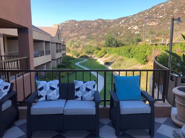 Outdoor patio area with black wicker seating including a loveseat and two chairs with cushions and patterned pillows. The patio overlooks a green lawn with a winding concrete path and hills in the background under a clear blue sky.