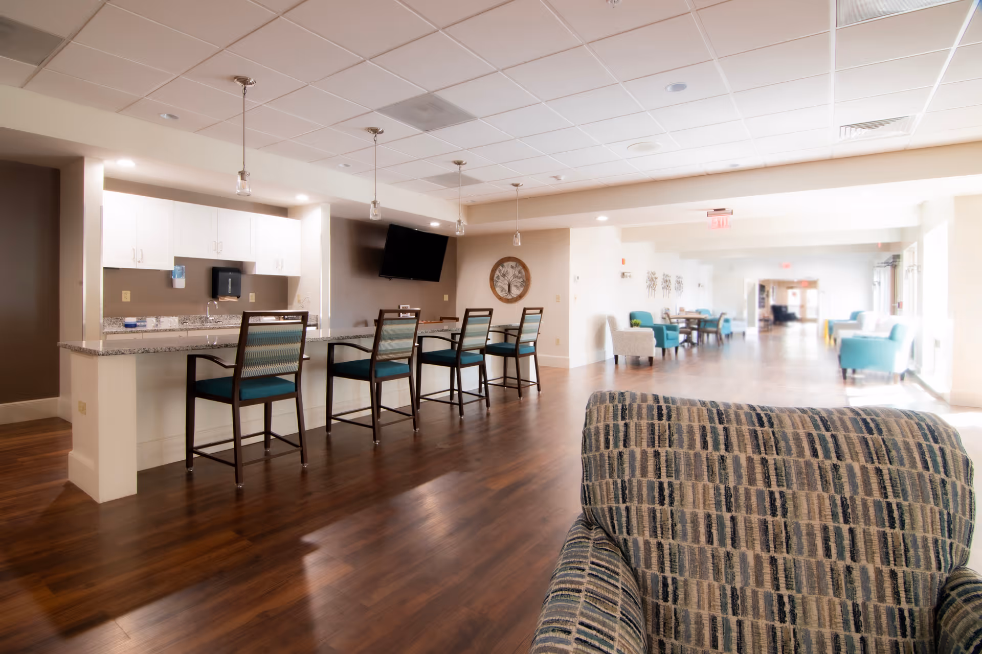 Bright communal lounge and kitchenette with a granite-topped bar and high chairs, seating area and TV in a senior living community.