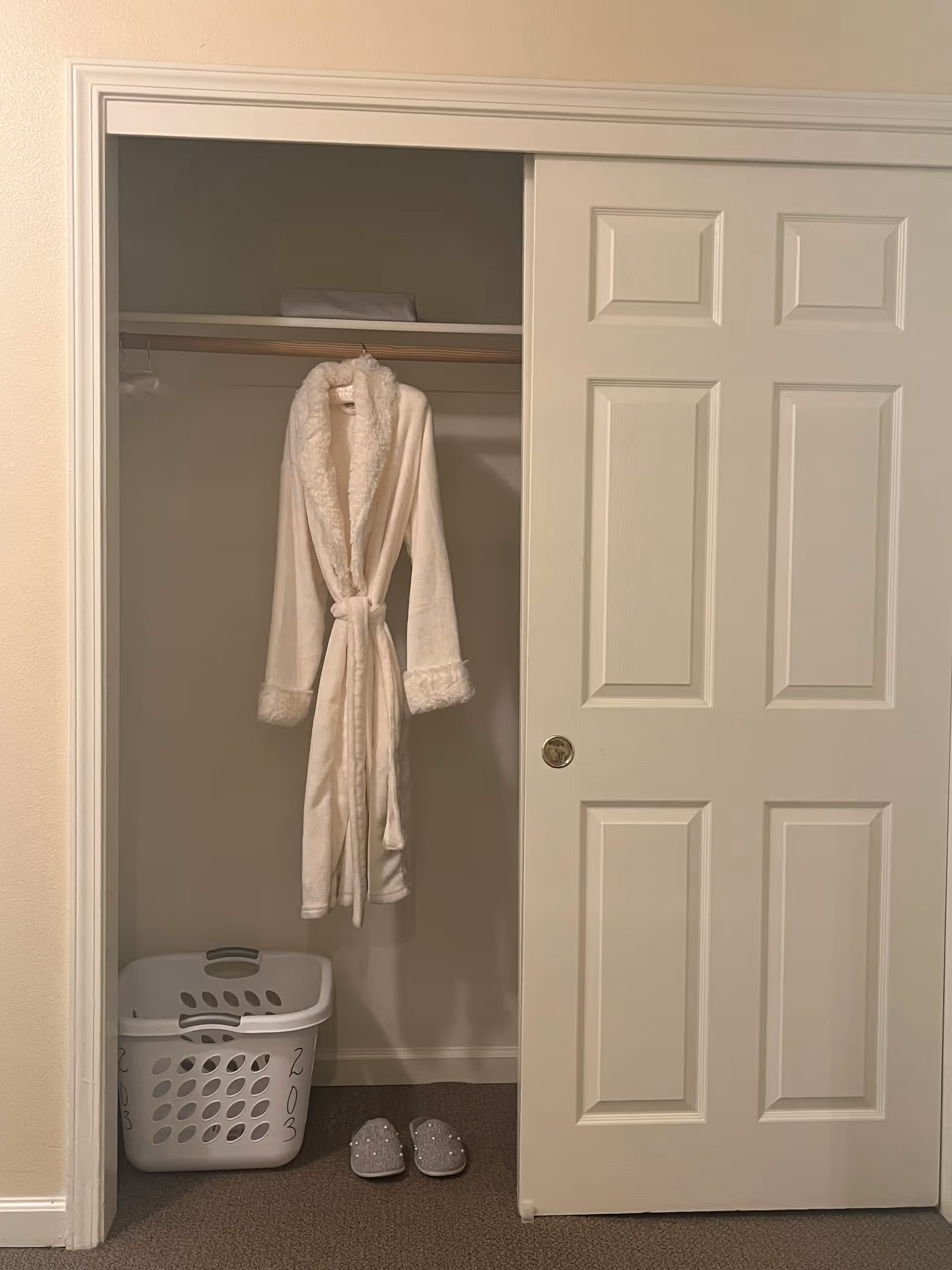 An open closet with a white sliding door. Inside the closet, a white bathrobe with a fluffy collar and cuffs is hanging on a wooden rod. Below the robe, there is a white laundry basket with the number 203 written on it and a pair of gray slippers with small decorative elements on the floor.