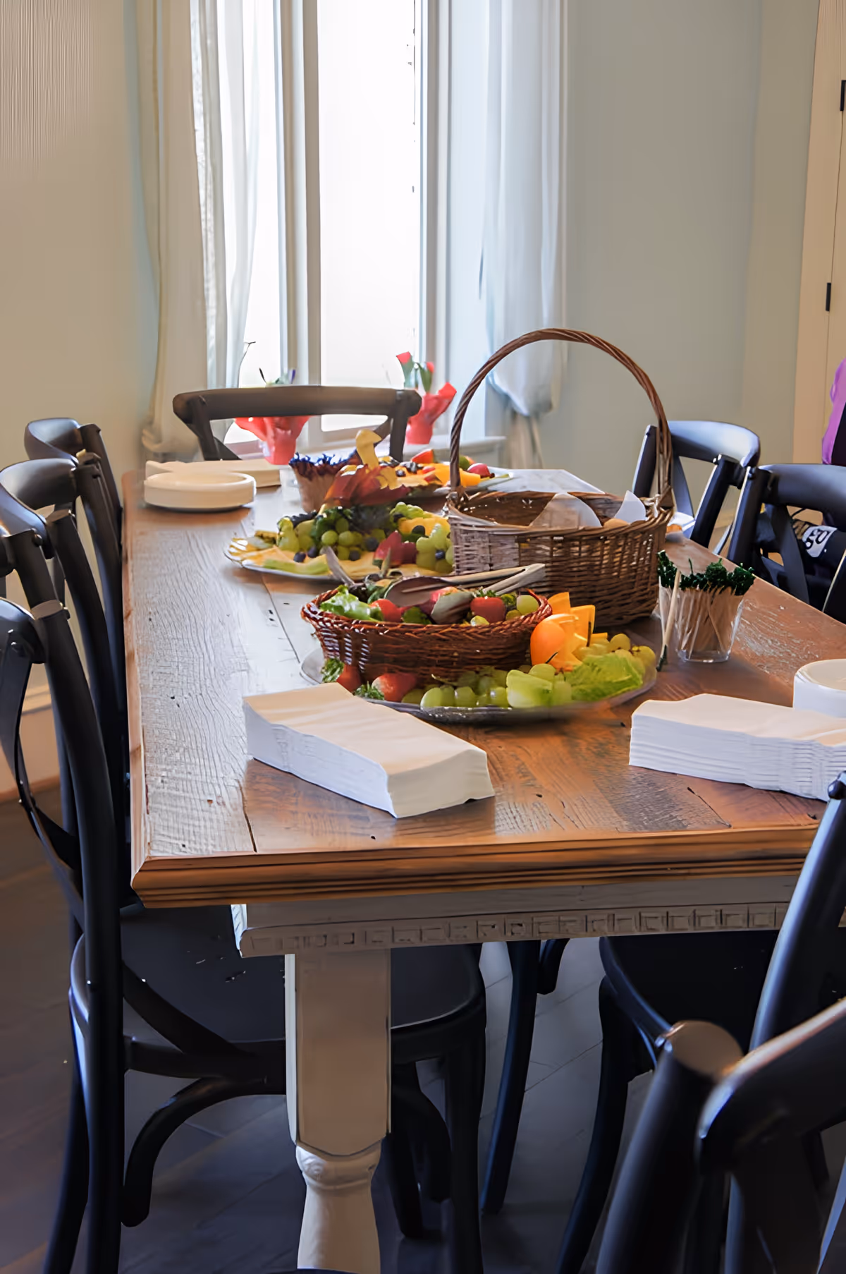A wooden dining table set with black chairs around it, featuring baskets and platters filled with fresh fruit, napkins, and toothpicks. The room has light-colored walls and a window with sheer white curtains in the background.