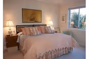 A cozy bedroom featuring a large bed with a pink and beige bedspread and multiple pillows. There are two bedside tables with lamps on each side of the bed. A framed artwork hangs above the headboard, and a window on the right side lets in natural light, showing some greenery outside.
