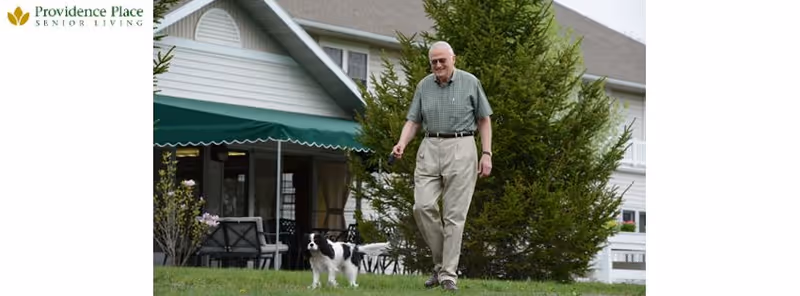 An elderly man walking a small black and white dog on a leash outside a senior living facility with a large green awning and white building in the background.