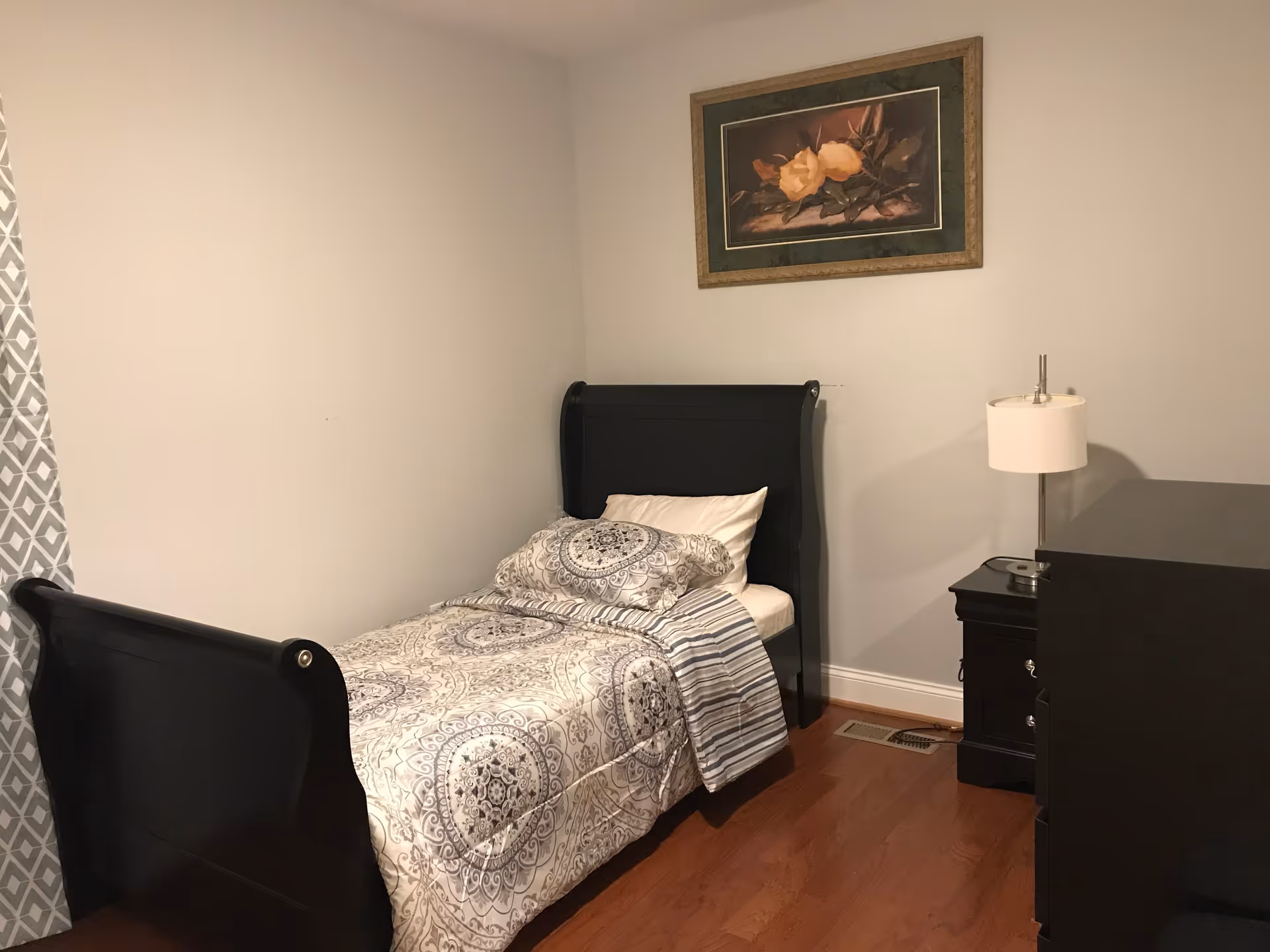 A small bedroom with a single black wooden bed featuring a patterned comforter and pillow. Next to the bed is a black nightstand with a white lamp. A black dresser is partially visible on the right side. The walls are light gray, and a framed painting of yellow flowers hangs above the bed. The floor is wooden, and a window with patterned curtains is on the left side.