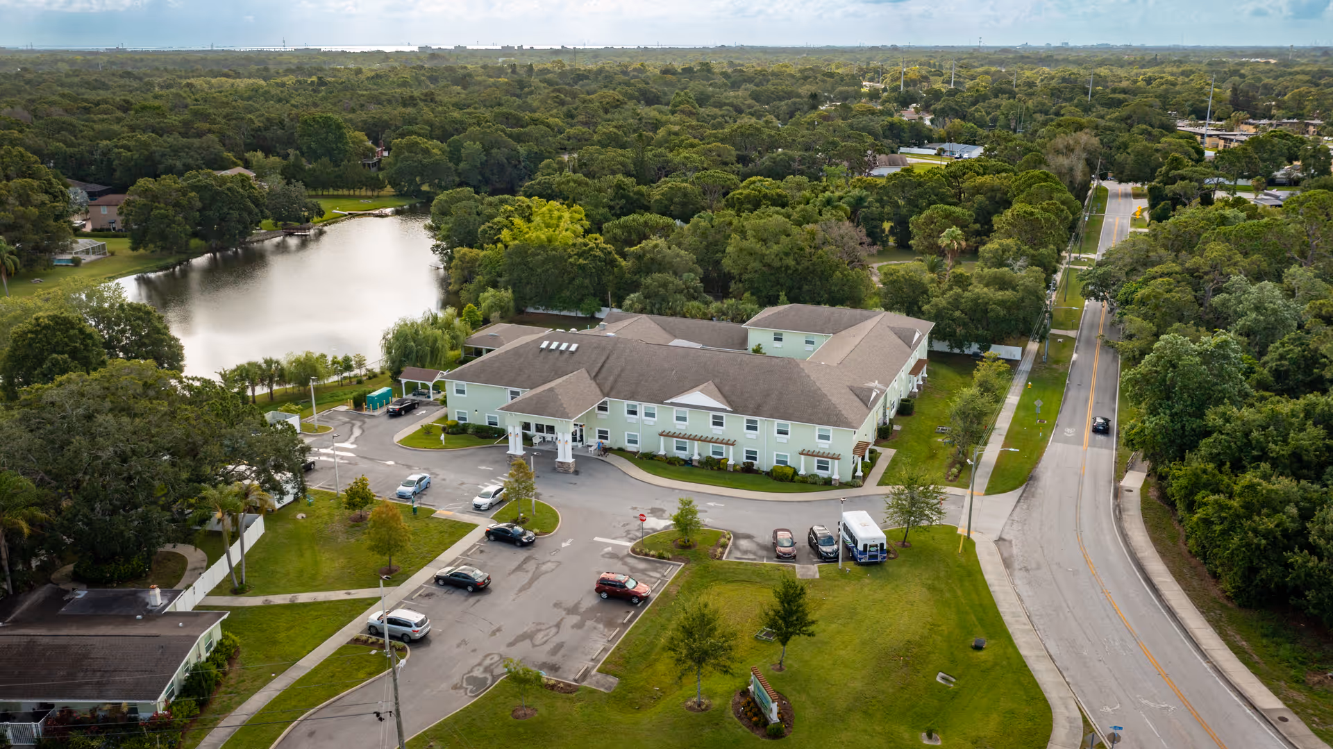 Aerial view of a senior living facility named Lake House surrounded by trees and greenery. The building is light green with a brown roof and has a parking lot with several cars. There is a pond nearby and a road running alongside the property.