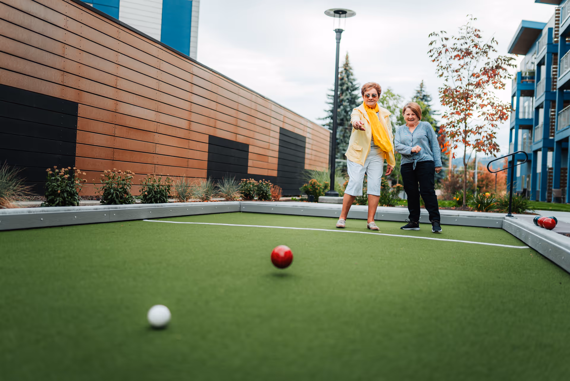 Two elderly women playing bocce ball on a green outdoor court next to a modern building with wooden paneling and blue accents, with plants and trees in the background.