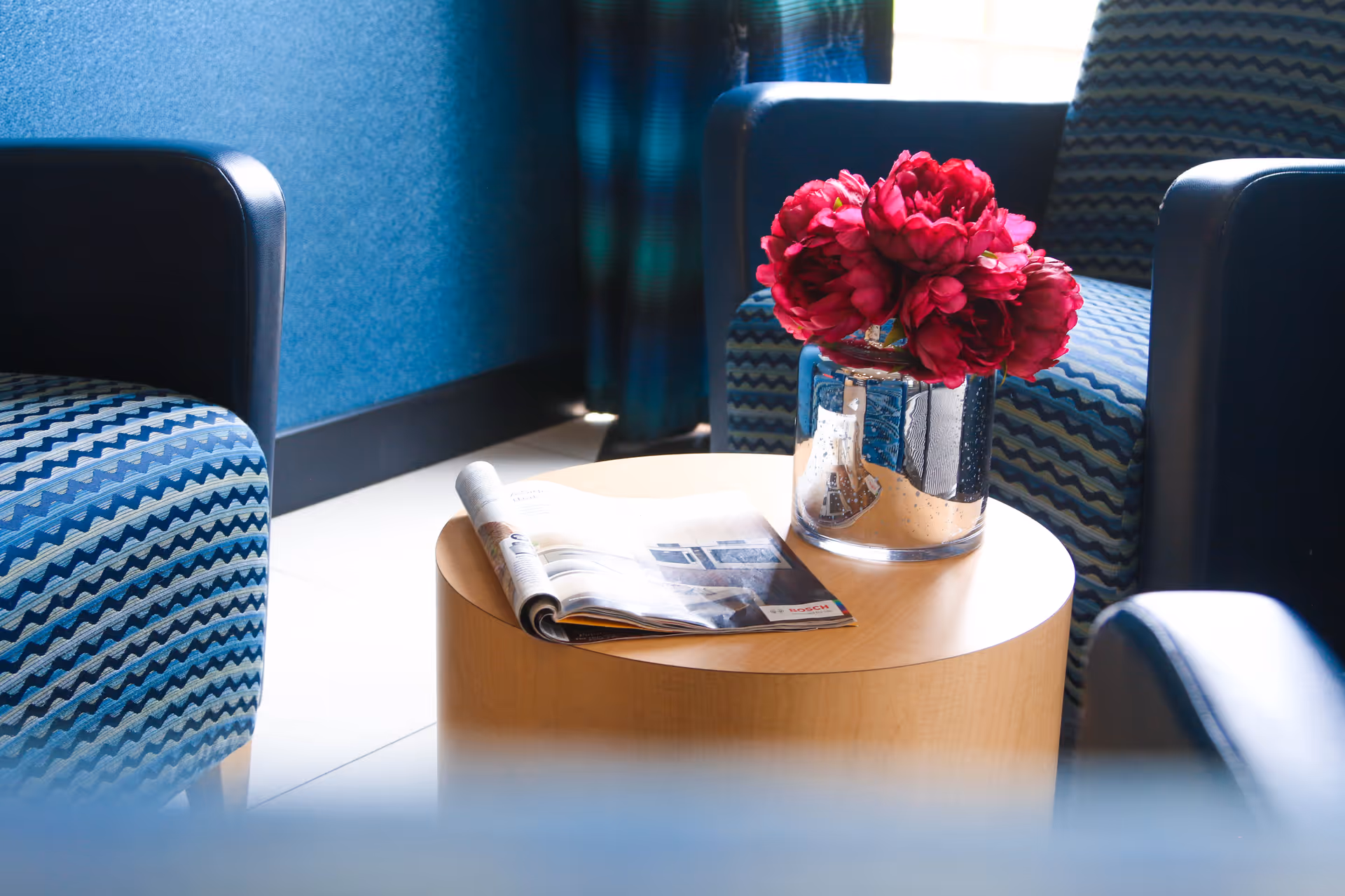 A cozy seating area with blue patterned armchairs surrounding a round wooden coffee table. On the table, there is a glass vase with vibrant red flowers and an open magazine.