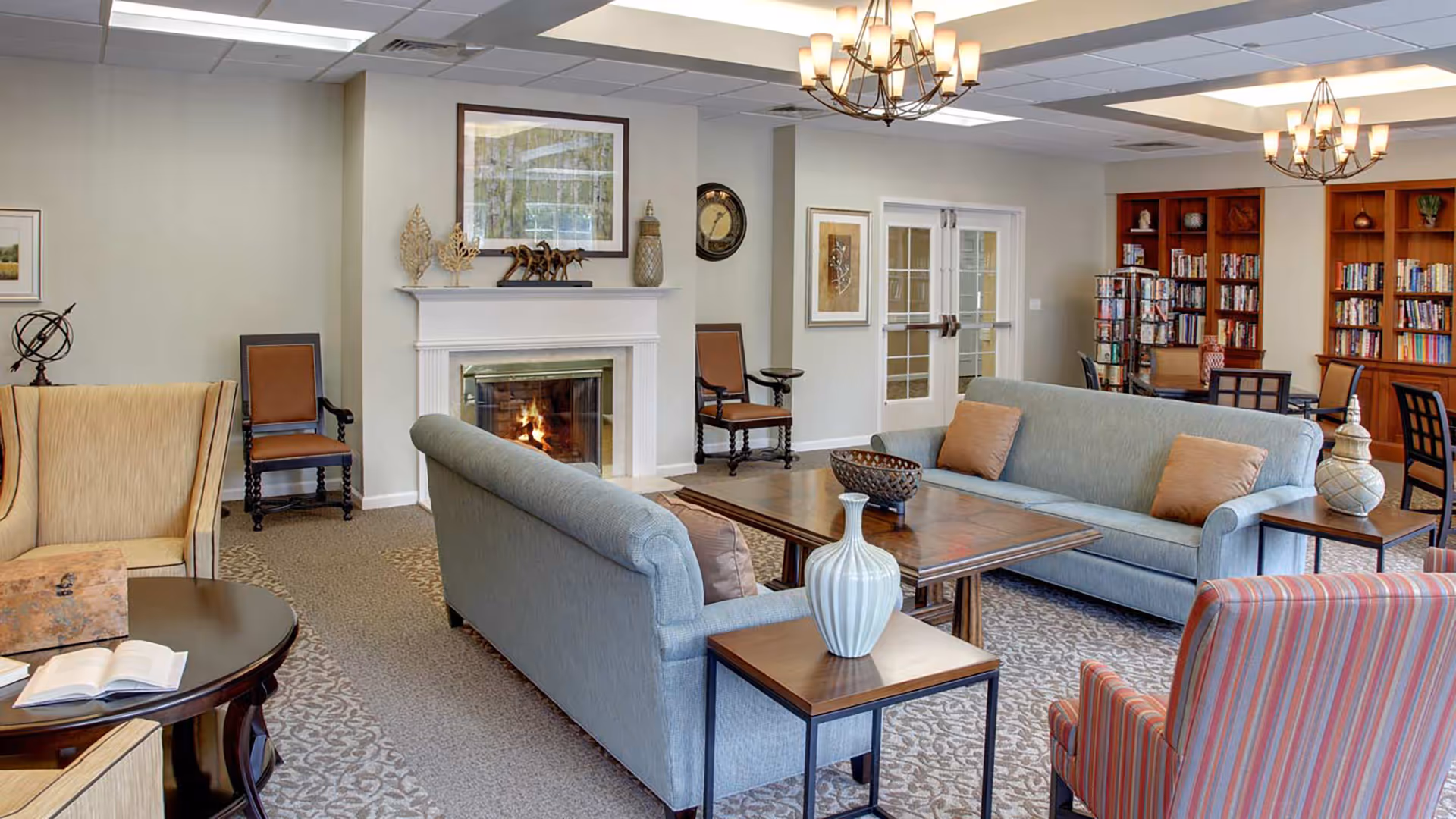 A cozy senior living facility common area with two blue sofas, a wooden coffee table, and side tables with decorative vases. There is a lit fireplace with a mantel decorated with art pieces and a framed picture above it. The room also features a beige armchair, a striped armchair, wooden chairs, a round table with an open book, and a bookshelf filled with books. Two chandeliers hang from the ceiling, and double glass doors are visible in the background.