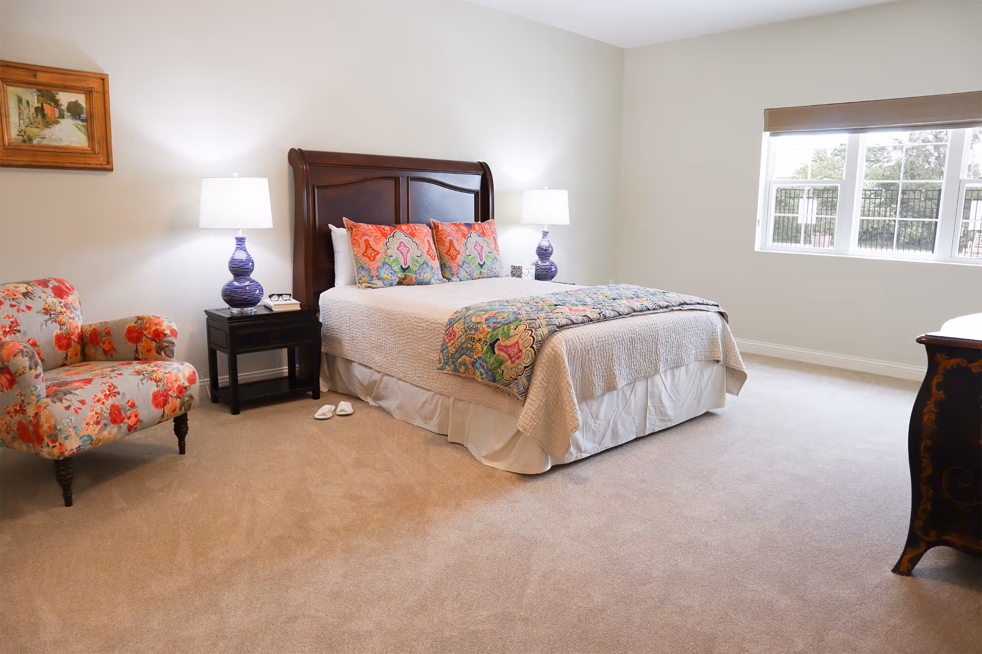 A bright and spacious bedroom with a large bed featuring a wooden headboard, colorful pillows, and a patterned throw blanket. There are two bedside tables with matching purple lamps, a floral upholstered armchair, a window with a view outside, and a framed painting on the wall.