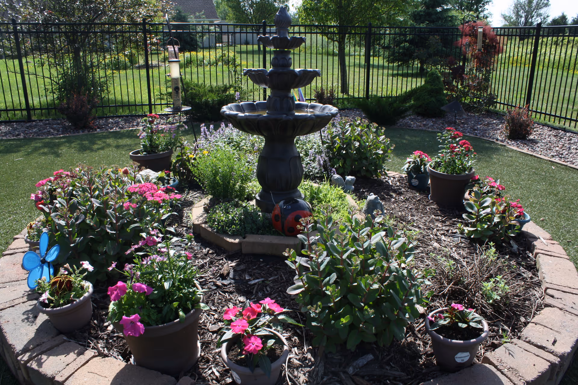 A circular garden bed with potted pink flowers and a three-tiered fountain in the center, surrounded by lawn and a metal fence.