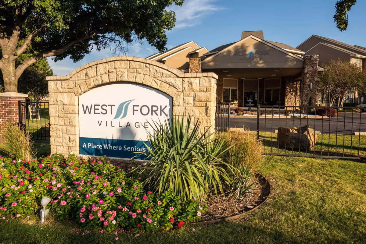 Stone sign for West Fork Village with landscaping including green plants and pink flowers in front, and the entrance to the facility visible in the background under a clear blue sky.