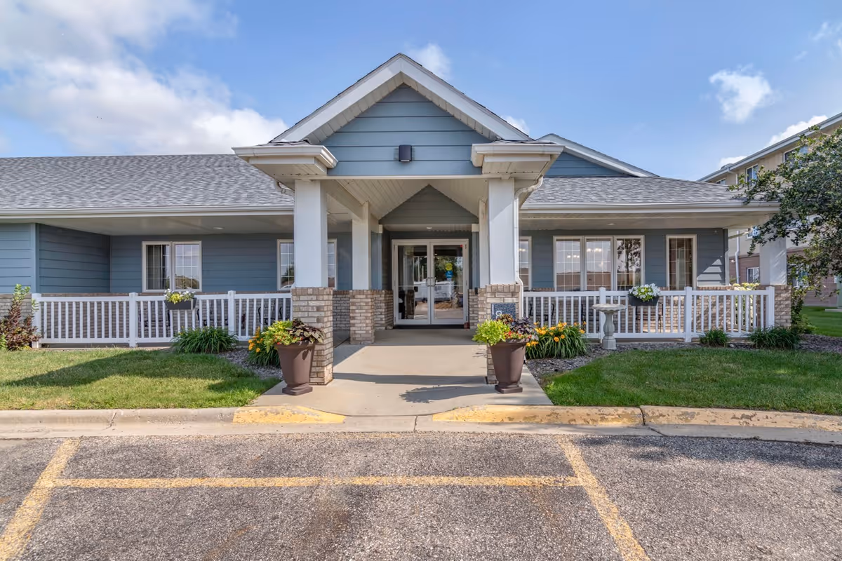 Front exterior view of Derian Place Senior Living building with a covered entrance, two large planters with flowers, a white railing, and green lawn on either side under a partly cloudy sky.