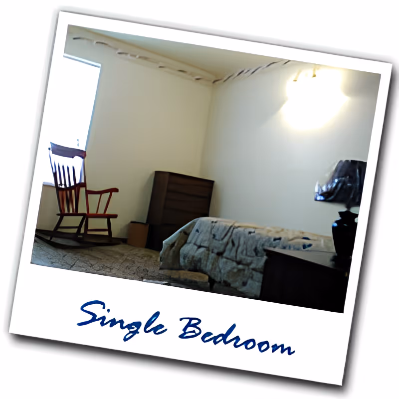 A simple single bedroom with a wooden rocking chair near a window, a dresser against the wall, a bed with a patterned blanket, and a nightstand with a lamp. The walls are plain and light-colored, and there is a ceiling light fixture.