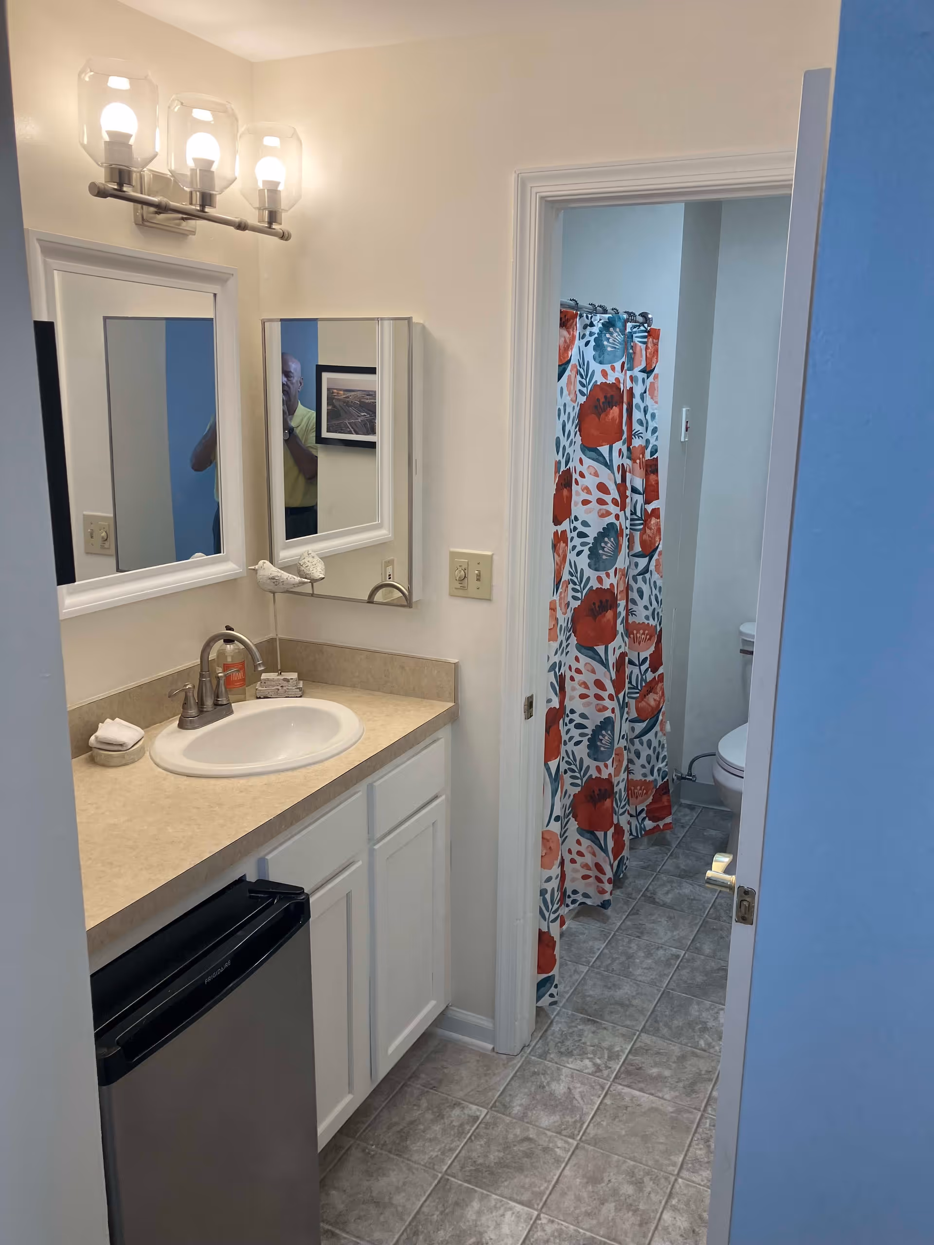 A bathroom area with a sink and countertop on the left, a mirror above the sink, and a light fixture with three bulbs. There is a small refrigerator under the counter. Through an open door, a shower with a colorful floral shower curtain and a toilet are visible. The floor is tiled in a gray pattern.