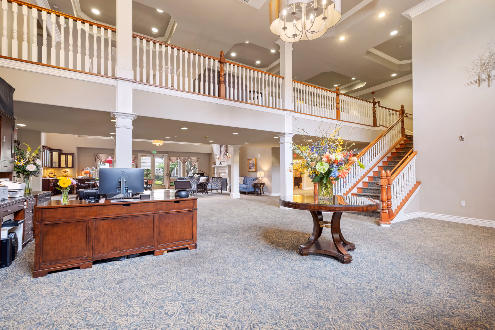 Spacious senior living facility lobby with a large wooden reception desk on the left, a round table with a colorful floral arrangement in the center, and a staircase with white railings and wooden banisters leading to an upper floor. The area is well-lit with recessed ceiling lights and a chandelier, featuring carpeted floors and neutral-colored walls.