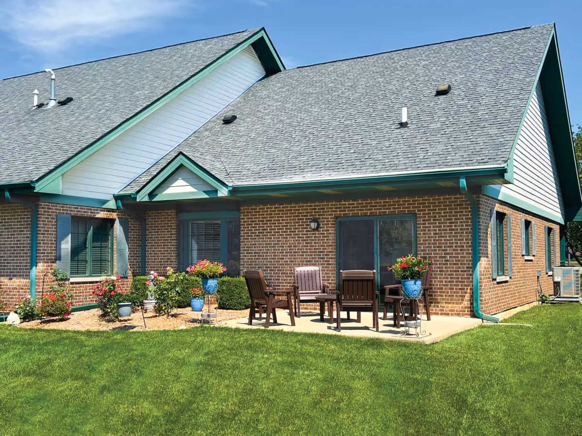 Exterior view of a brick senior living facility building with a gray shingled roof and green trim. There is a small patio with four wooden chairs and a table, surrounded by potted plants and flowers. The lawn is green and well-maintained under a clear blue sky.