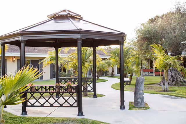 Outdoor courtyard featuring a metal gazebo, paved walkways, palm trees, and a single-story building in the background.