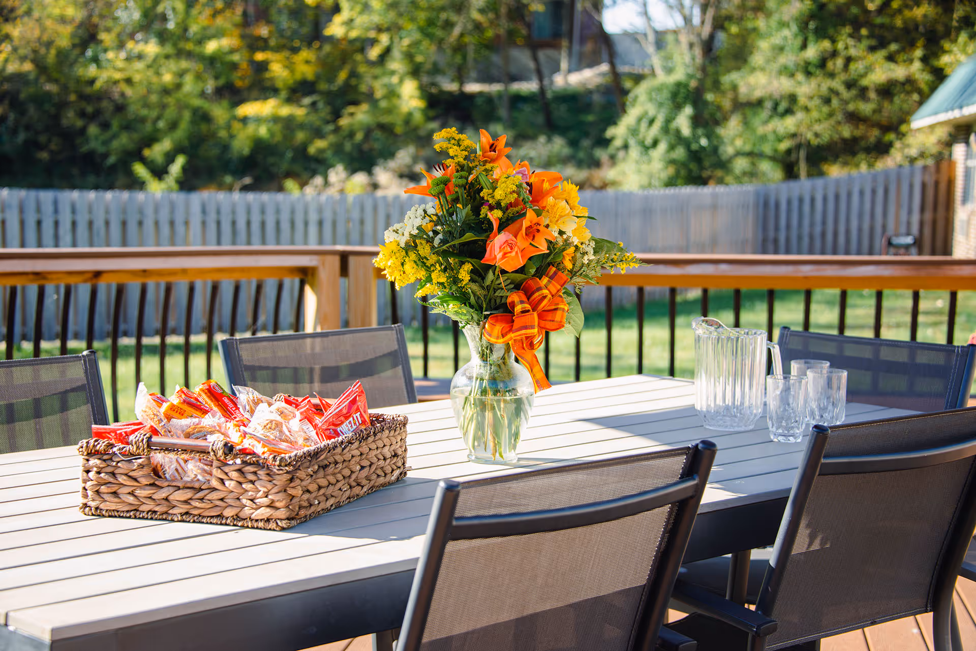 Outdoor patio table with a basket of individually wrapped snacks, a clear glass vase with a colorful bouquet of orange, yellow, and white flowers tied with an orange ribbon, and a clear pitcher with matching glasses. The table is surrounded by black mesh chairs and is set on a wooden deck with a wooden railing and a fenced backyard with trees in the background.