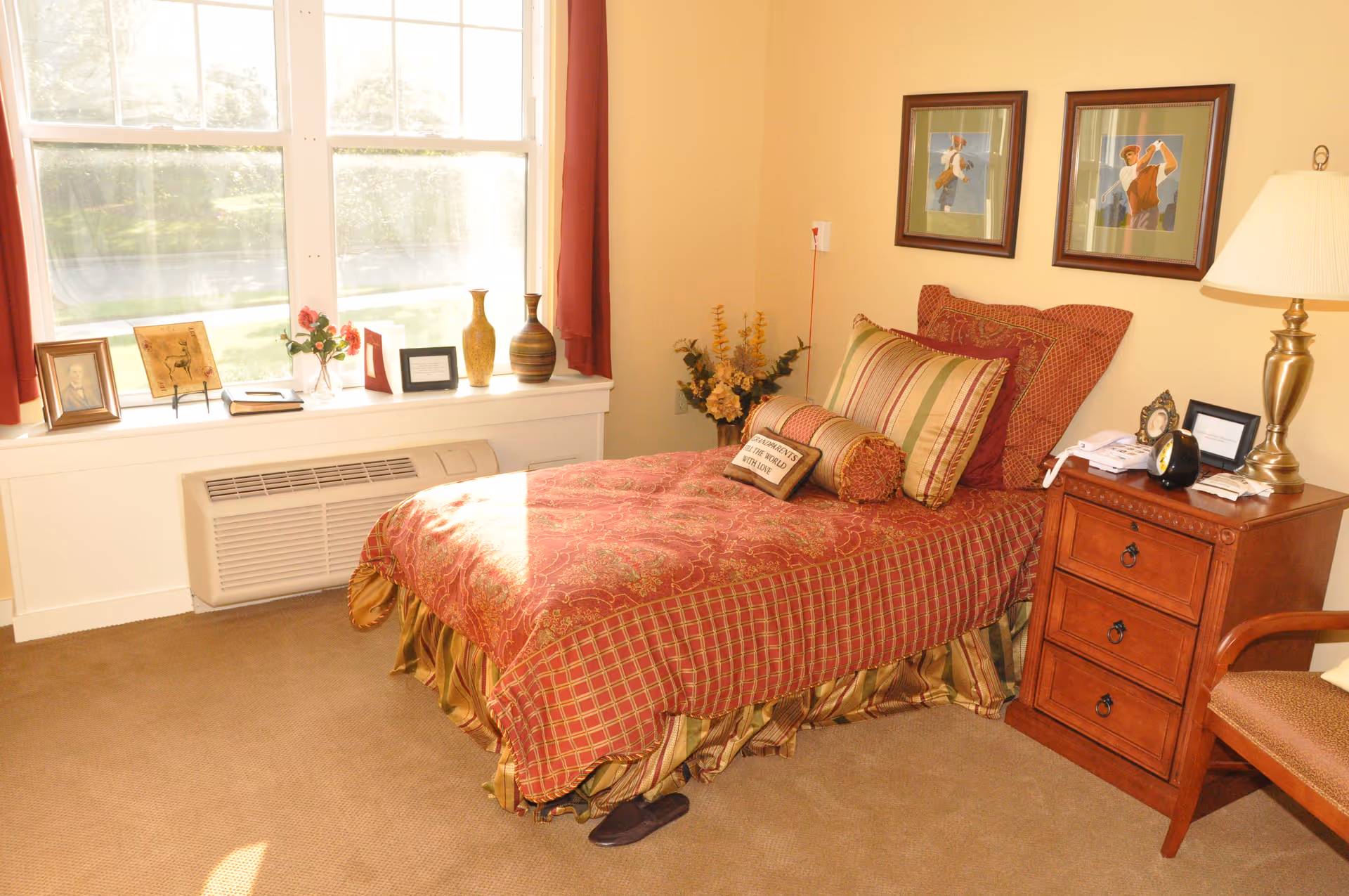 Sunlit bedroom with a single bed in red patterned bedding, a wooden nightstand, large window with decor on the sill, and framed pictures on the wall.