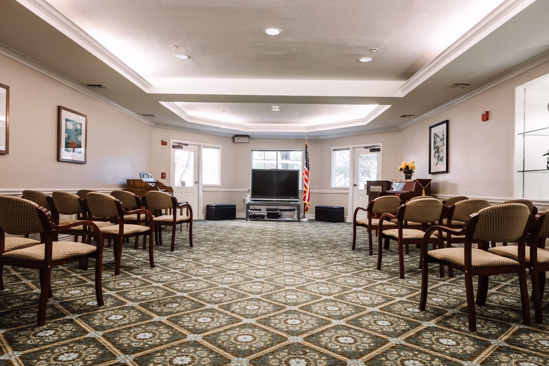 A spacious room with patterned carpet and two rows of wooden chairs with cushioned seats facing a television on a stand. The room has beige walls with framed artwork, a piano with a flower vase, and an American flag near the TV. There are two doors and a window behind the TV, and the ceiling has recessed lighting with a tray ceiling design.