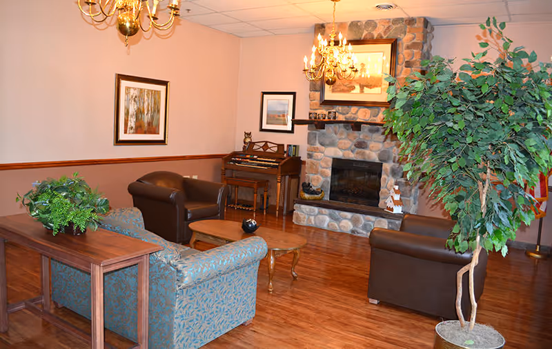 A cozy common room with sofas, armchairs, a coffee table, a stone fireplace, chandeliers, and potted plants.