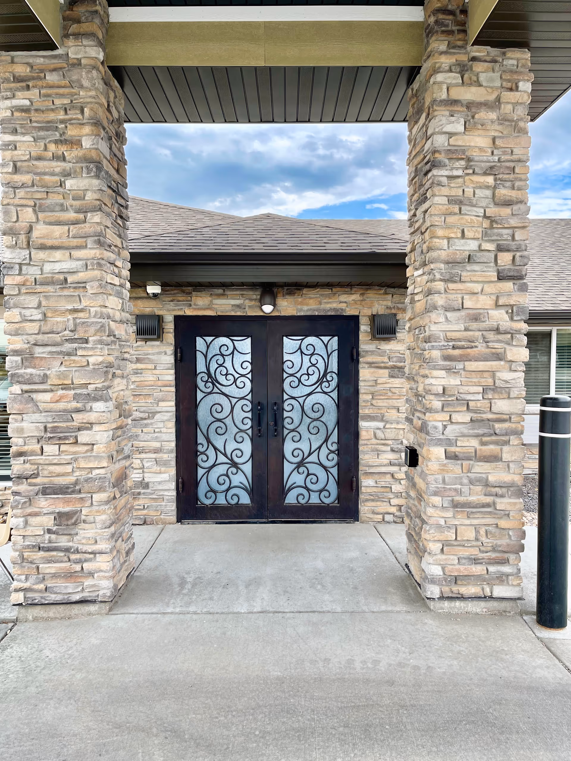 Entrance to a building with double black metal doors featuring decorative scrollwork glass panels, flanked by two large stone pillars under a covered porch with a concrete walkway leading up to the doors.
