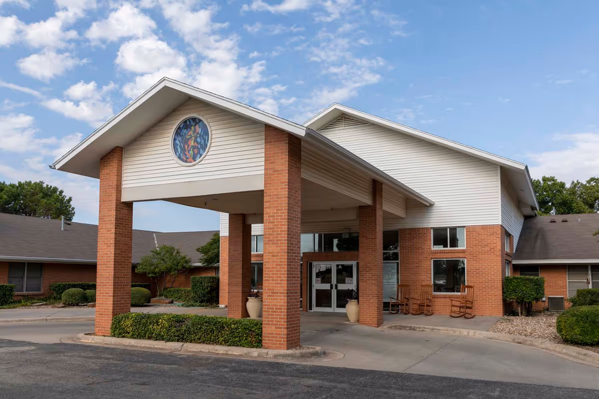 Exterior view of Wisteria Place Assisted Living facility showing a covered entrance with brick pillars, a circular stained glass window above the entrance, and surrounding landscaping with bushes and trees under a partly cloudy sky.