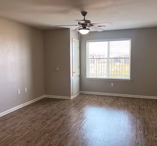 Empty sunlit living room with wood-look flooring, a ceiling fan, and a large window with blinds.
