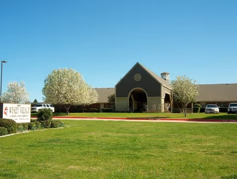 Front exterior of Wesley Village senior living building with a large grassy lawn, blossoming trees, and a driveway with parked cars.