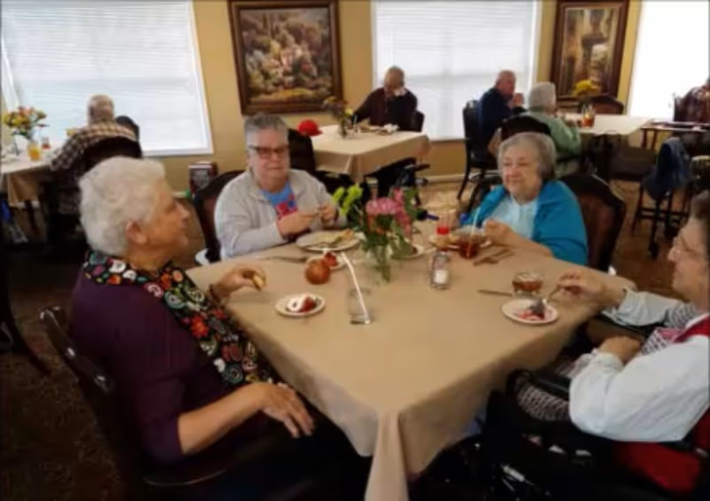 Four elderly residents sit around a dining table in a communal dining room enjoying dessert.