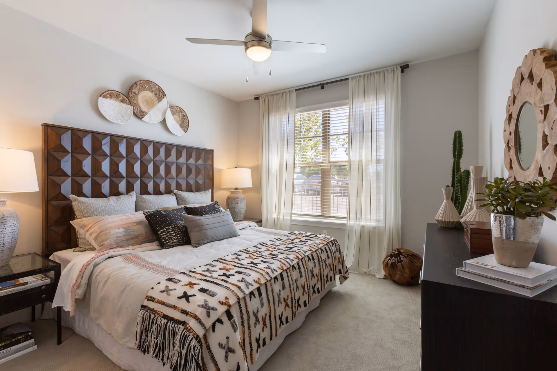 A cozy bedroom with a large wooden headboard bed adorned with multiple pillows and a patterned throw blanket. There are two bedside tables with lamps, a window with sheer curtains letting in natural light, a dresser with decorative items including a plant and a round wooden mirror on the wall, and a ceiling fan with a light.