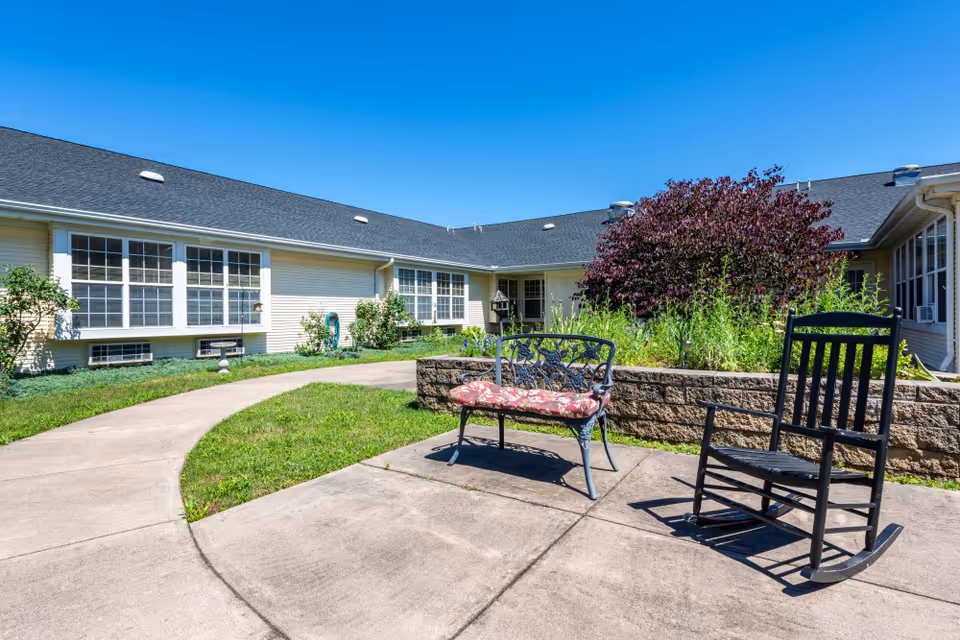 Outdoor courtyard area at American House Johnson City featuring a concrete pathway, a black metal bench with a floral cushion, a black wooden rocking chair, and a raised garden bed with green plants and a purple-leafed tree under a clear blue sky.