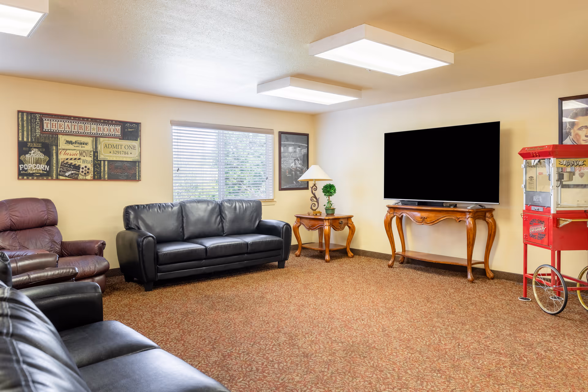 A cozy senior living community lounge with a black leather sofa and armchairs, a wooden side table with a lamp and small plant, a large flat-screen TV on a wooden console table, a red popcorn machine, and wall art including a theatre-themed poster and a black and white photo. The room has beige walls, a window with blinds, and patterned carpet flooring.