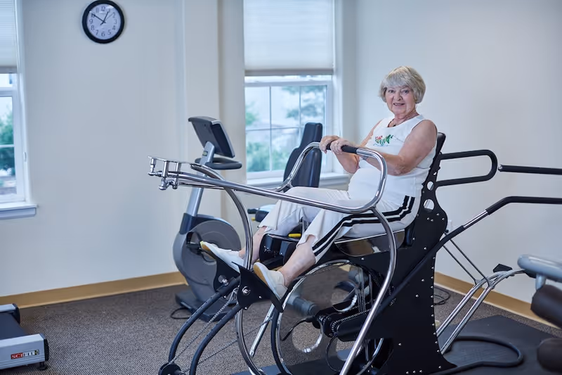 An elderly woman with gray hair wearing a white sleeveless top and white pants with black stripes is seated on a piece of exercise equipment in a fitness room. The room has two windows with white blinds, a wall clock showing 9:10, and other exercise machines in the background.
