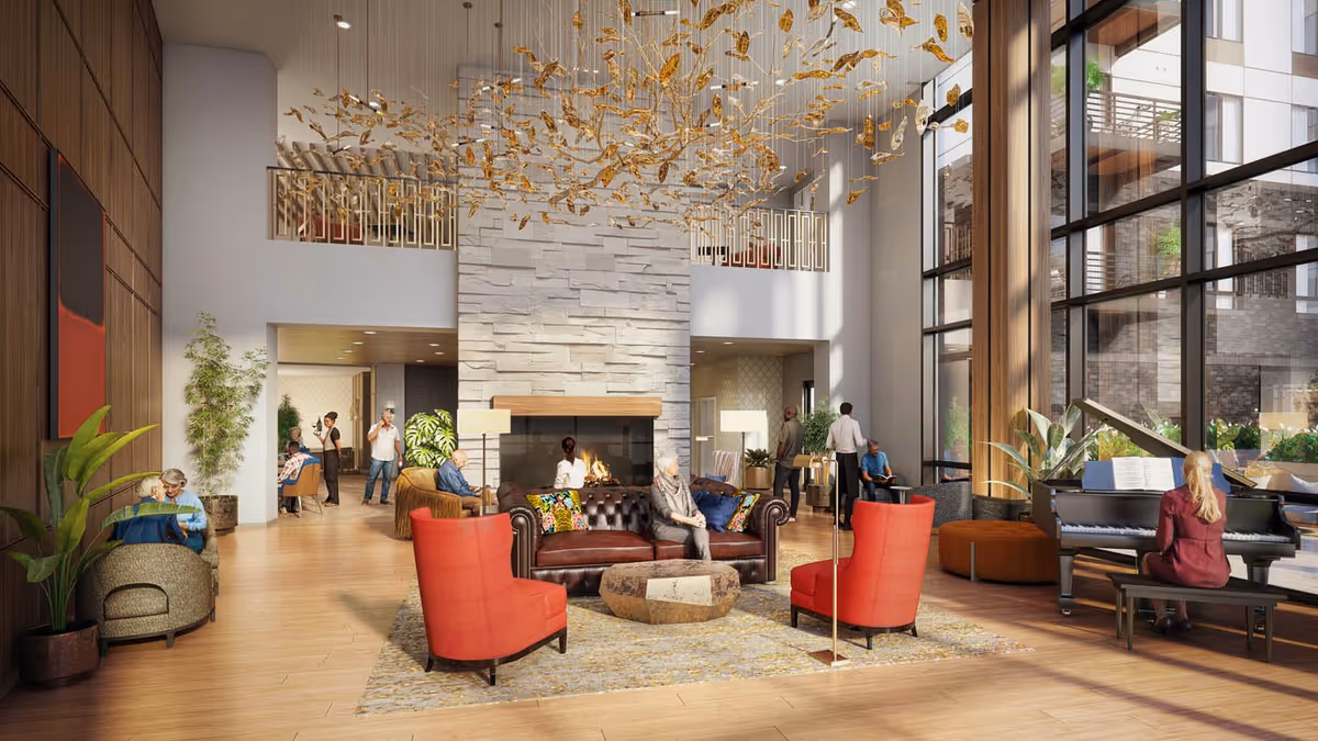 A spacious and well-lit senior living facility common area with a large stone fireplace in the center. The room features a brown leather sofa with colorful pillows, two red armchairs, and a unique stone coffee table on a patterned rug. A woman is playing a grand piano near tall windows that let in natural light. Several seniors are seated or standing, engaging in conversation or relaxing. The ceiling has an artistic gold hanging installation, and there are plants and modern decor throughout the space.