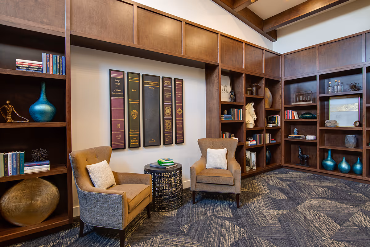 A cozy reading nook in a senior living facility featuring two cushioned armchairs with pillows, a small round side table with books, and built-in wooden bookshelves filled with books and decorative items. The walls have large book spine art pieces, and the carpet has a geometric pattern.