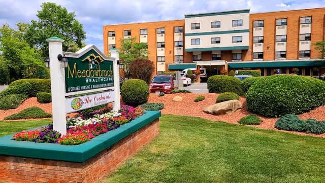 Front entrance of Meadowbrook Healthcare with a landscaped sign, flowerbeds, and the multi-story brick facility in the background.