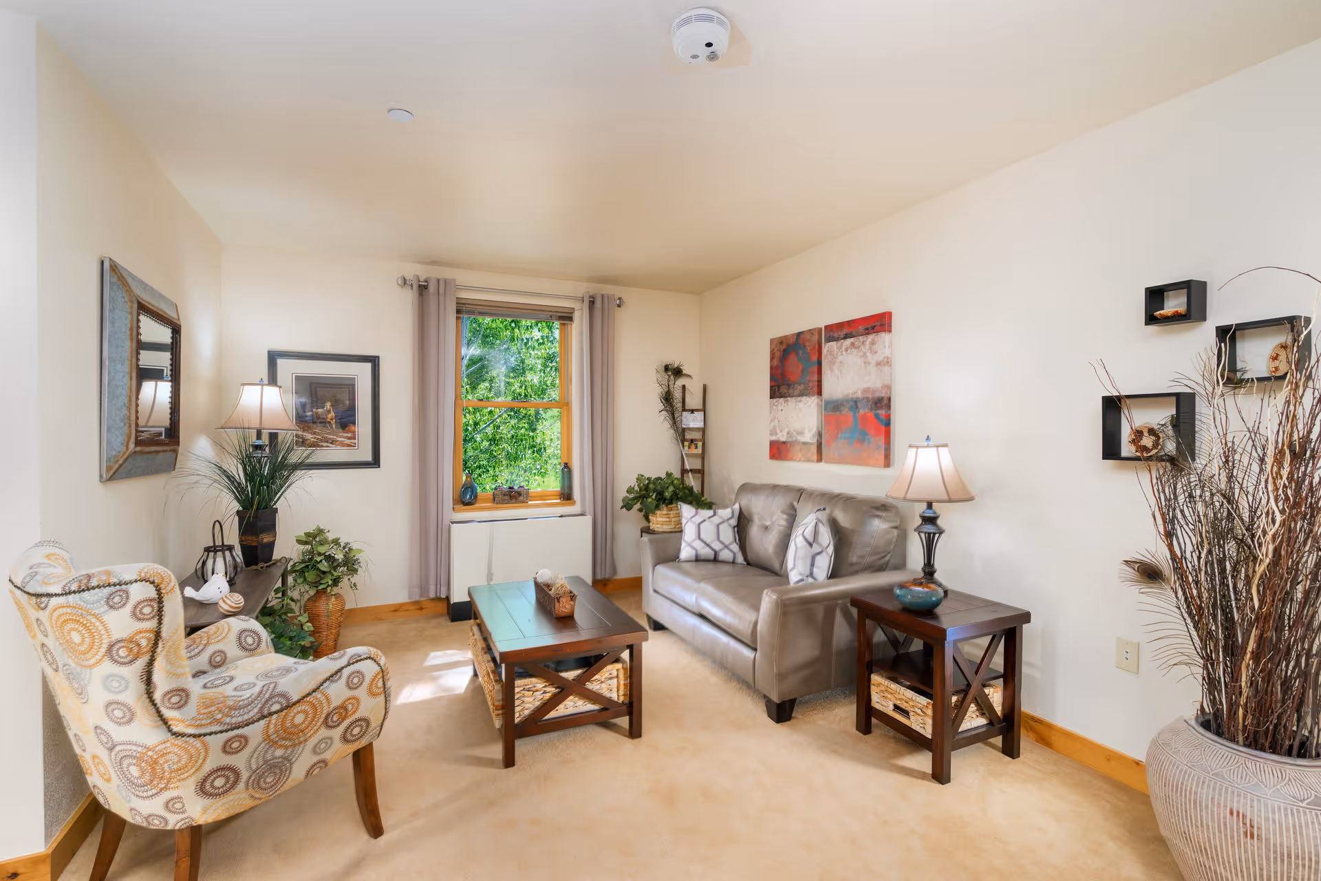 Well-lit cozy living room with a sofa, patterned armchair, coffee table, lamps, artwork, and plants.
