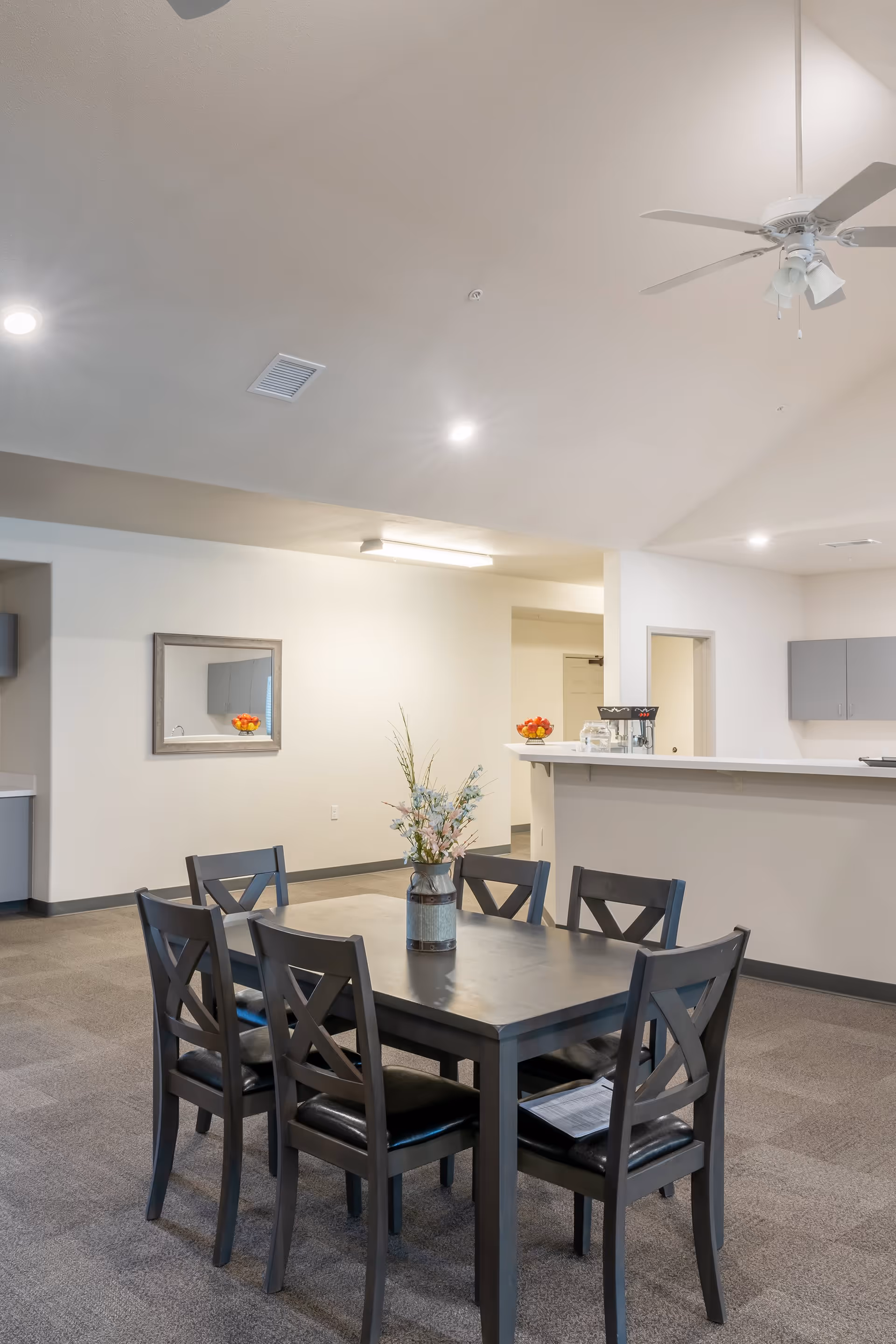 A dining area with a rectangular dark wooden table surrounded by six matching chairs with black cushioned seats. A vase with flowers is placed in the center of the table. In the background, there is a counter with a bowl of fruit and some kitchen appliances. The room has beige walls, carpeted flooring, a ceiling fan, and recessed lighting.