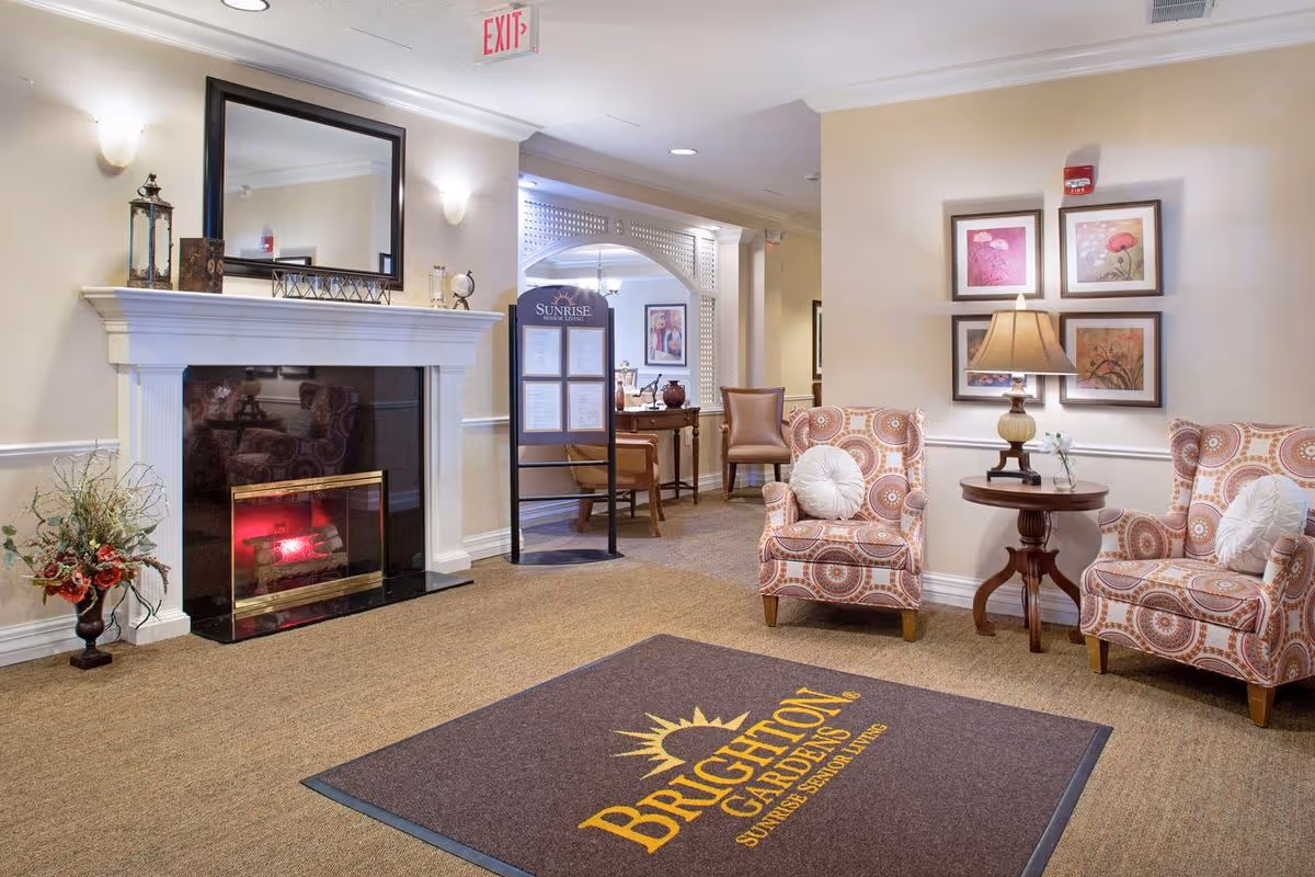 A cozy seating area in a senior living facility featuring two patterned armchairs with white round pillows, a wooden side table with a lamp, and framed floral artwork on the wall. There is a fireplace with a large mirror above it, decorative items on the mantel, and a floral arrangement on the floor. A carpet with the Brighton Gardens logo is placed on the floor, and a hallway with additional seating and a signboard is visible in the background.