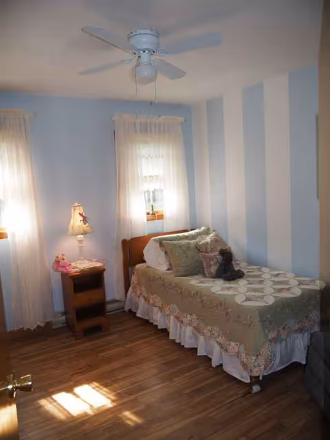 Cozy bedroom with a single bed, nightstand lamp, blue striped walls, curtains, and a ceiling fan.